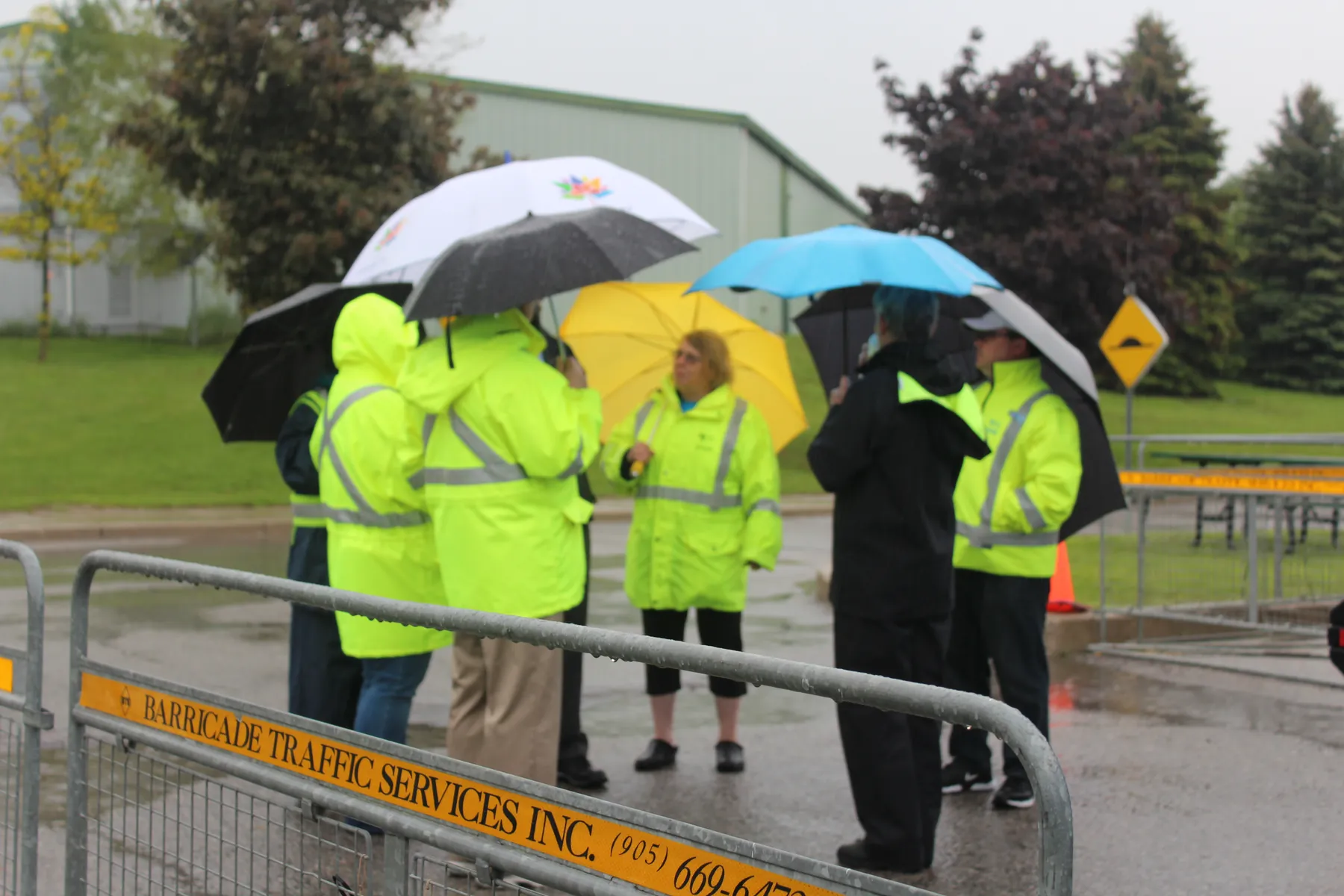 Roadeo participants in high-visibility jackets under umbrellas beside a course barricade.